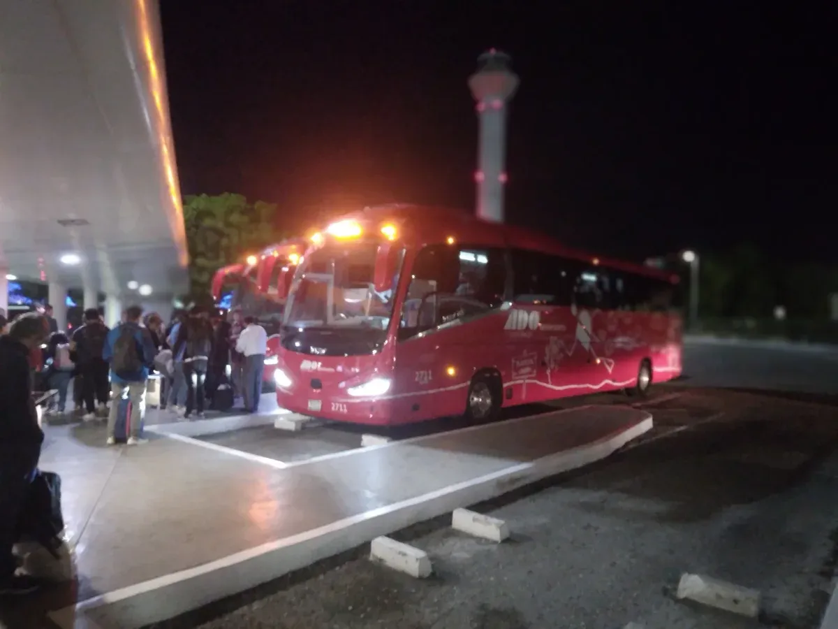 Red coach bus at night at the Terminal 2 platform with passengers queuing and the control tower behind