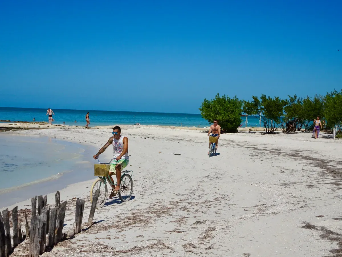 Two cyclists riding along a white sand beach on Holbox with calm turquoise water beyond